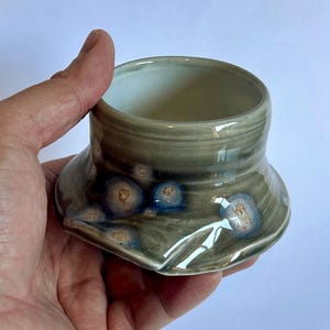 May include: A small, round ceramic bowl with a flared base and a smooth, glossy finish. The glaze is a muted sage green with distinct, circular crystalline patterns in shades of deep blue and warm tan scattered across the exterior surface.