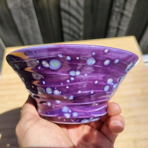 May include: A small, flared ceramic bowl with a glossy purple glaze featuring a swirling texture and scattered light blue and white crystalline spots across the exterior surface, creating a speckled appearance on the rounded vessel.