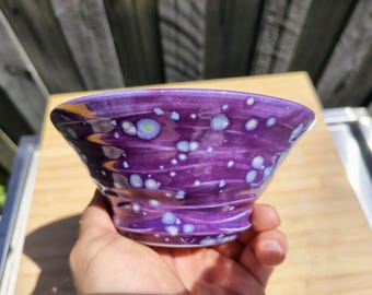 Peut inclure: Un petit bol en céramique évasé avec un vernis violet brillant présentant une texture tourbillonnante et des taches cristallines bleu clair et blanches dispersées sur la surface extérieure, créant un aspect moucheté.