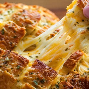 May include: A pull-apart loaf of golden-brown bread with deep cuts, filled with melted white and orange cheese and sprinkled with green herbs. A piece is being pulled away, showing long, stretchy strands of melted cheese. Text reads The Sugared Hour.