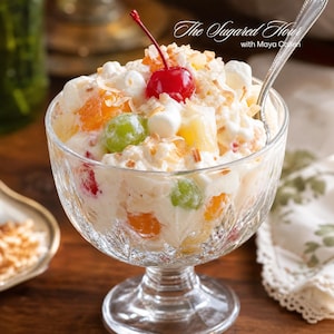 Könnte beinhalten: Eine gläserne Dessertschale gefüllt mit Ambrosia-Salat aus Mandarinen, grünen Trauben, Ananasstücken, weißen Marshmallows und Kokosraspeln, garniert mit einer roten Kirsche und einem Silberlöffel. Der Text lautet The Sugared Hour with Maya Callen.