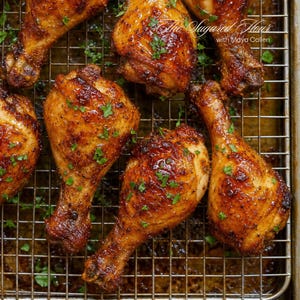 May include: Roasted chicken drumsticks with a golden-brown glaze and a light dusting of fresh green parsley, arranged on a metal wire cooling rack. The text "The Sugared Flour with Maya Callen" is visible in the upper right corner of the image.