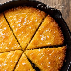 Pode incluir: Um pão de milho dourado cortado em fatias triangulares dentro de uma frigideira de ferro fundido preta. A superfície está envidraçada com um líquido brilhante e coberta com cristais de sal branco grosso. O texto "The Sugared Flour with Maya Callen" é visível.