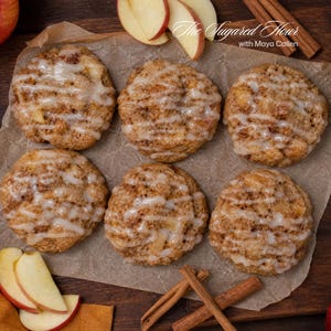 May include: Six round, golden-brown apple cinnamon cookies arranged on parchment paper, each drizzled with a white sugar glaze. The text "The Sugared Flour with Maya Callen" appears in the upper right corner, accompanied by apple slices and cinnamon sticks.
