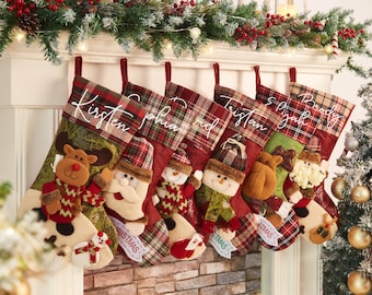 Op de afbeelding: Zes kerstsokken hangen op een rij, elk met een geruite boord en een wit geborduurde naam. De sokken zijn voorzien van pluche applicaties van een rendier, de kerstman en sneeuwpoppen, in de kleuren rood, groen en bruin.