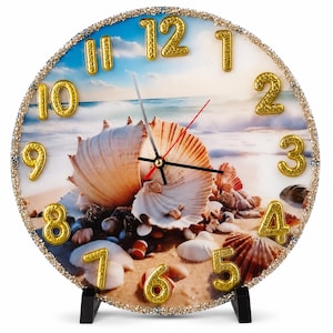 May include: A round wall clock featuring a beach scene with seashells on sand and a blue ocean horizon. The clock face has raised gold-colored numbers and a border encrusted with small gold beads. It includes black hour and minute hands and a red second hand.