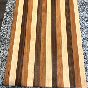 May include: A rectangular wooden cutting board featuring a vertical striped pattern. The board is constructed from alternating planks of light cream, medium tan, and dark brown wood, creating a multi-tonal appearance across the entire surface.