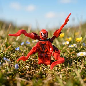 Puede incluir: Una minifigura de plástico roja con un patrón de araña negro en el torso y las piernas, una cara sonriente con ojos blancos y cuatro apéndices rojos en forma de tentáculo que salen de la espalda, de pie en un campo de hierba y flores.