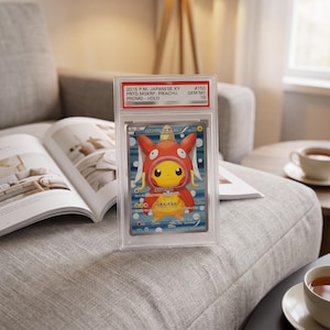 May include: A 2015 Japanese XY Promo Holo Pikachu trading card encapsulated in a clear protective slab. The card features Pikachu wearing a red Magikarp costume against a blue background with white circles. The label reads 2015 P.M. JAPANESE XY PRTD.MGKRP. PIKACHU PROMO-HOLO GEM MT 10.