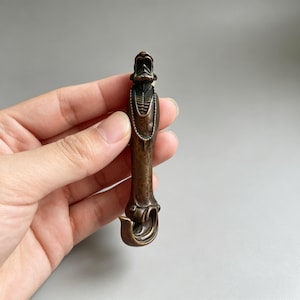 May include: A bronze-toned metal object shaped like a stylized figure with an open-mouthed head at the top, a beaded necklace detail, and a curved hook at the base. The vertical piece has a dark, weathered finish with visible metallic highlights.