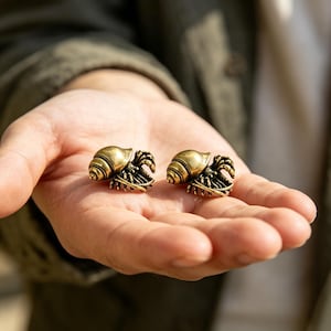 May include: Two small, metallic gold-toned hermit crab figurines. Each piece features a detailed spiral shell and articulated legs, crafted with a polished finish that reflects light. These miniature sculptures capture the form of a crustacean.