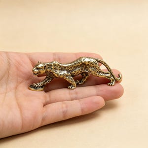 Könnte beinhalten: Eine kleine, goldfarbene Leopardenfigur aus Metall in einer schleichenden Haltung. Der Leopard weist detaillierte Flecken auf seinem Körper auf und ist so dargestellt, dass eine Vorderpfote auf einem kleinen, runden, münzähnlichen Objekt ruht.