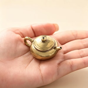 Op de afbeelding: Een miniatuur theepot van messing met een rond lichaam, een klein gebogen handvat, een korte tuit en een deksel met een centrale knop. Het metalen oppervlak heeft een gepolijste, goudgele afwerking die licht reflecteert op de gladde buitenkant.