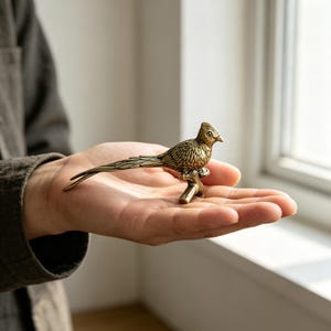 以下が含まれることがあります： 枝にとまった長い尾を持つ鳥のアンティーク調の小さな真鍮製フィギュア。金属製の彫刻には、詳細な羽の質感、頭部の目立つトサカ、そして体から水平に伸びる細く長い尾が表現されています。