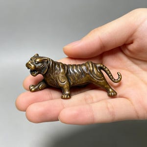 May include: A small, solid bronze-colored metal figurine of a tiger in a prowling stance. The tiger features detailed engraved stripes across its body, a textured surface, an open mouth showing teeth, and a curved tail extending upward.
