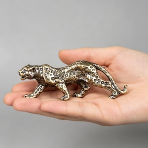 May include: A small, metallic bronze-toned leopard figurine in a prowling stance. The sculpture features a detailed, textured coat with dark, recessed spots across its body, a long curved tail, and an open mouth showing teeth.
