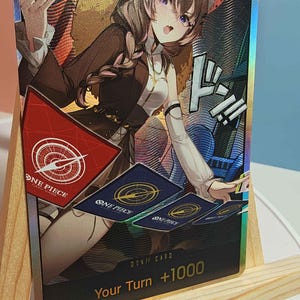 Puede incluir: Una carta coleccionable holográfica de One Piece con un personaje de anime de cabello castaño y orejas de gato. La carta muestra el texto DON!! CARD y Your Turn +1000, junto con varias ilustraciones de cartas y un emblema triangular rojo.