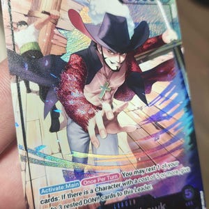 Peut inclure: Une carte à collectionner One Piece holographique représentant Dracule Mihawk avec une puissance de 5000. La carte montre le personnage portant un chapeau noir et un manteau rouge, avec le texte détaillant la capacité Activate:Main.