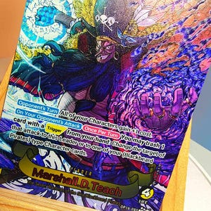 Könnte beinhalten: Eine One Piece Sammelkarte mit Marshall D. Teach, auch bekannt als Blackbeard, mit holografischem Finish. Die Karte zeigt die Figur mit Piratenhut, dunklem Haar und violetten Energieeffekten sowie Spieltext und einem Lebenswert von 4.
