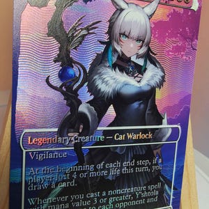 Puede incluir: Una carta coleccionable holográfica titulada Y'shtola, Night's Blessed, que muestra a un personaje brujo gato de pelo blanco sosteniendo un bastón oscuro con un orbe azul. La carta muestra texto del juego, símbolos de maná y un valor de 2/4.