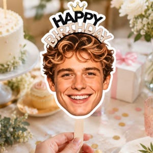 May include: A custom birthday cake topper featuring a cutout of a smiling person with wavy brown hair. The topper includes a gold crown icon at the top and the text HAPPY BIRTHDAY in black and glittery gold lettering on a white background.