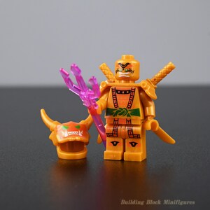 Peut inclure: Une figurine de bloc de construction en plastique jaune avec un visage imprimé aux sourcils noirs et dents blanches. Elle porte une armure orange avec une ceinture verte, tient un trident violet translucide et possède un masque cornu orange.