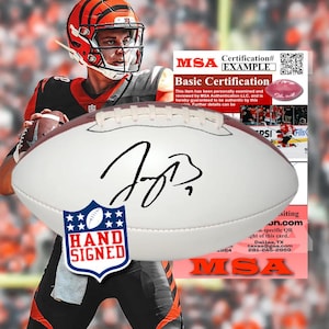 Op de afbeelding: Een witte lederen American football met een handtekening in zwarte inkt in het midden. Een blauwe schildvormige sticker met witte sterren en de tekst HAND SIGNED is onderaan bevestigd. Een MSA-certificeringskaart met tekst is zichtbaar.