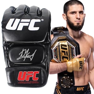 May include: A black UFC branded MMA glove featuring a white logo, a silver signature, and a red and white UFC logo on the wrist strap. The strap includes the text Fight the Passion Conquer the Moment. A gold glove and championship belt are visible.