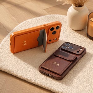 Peut inclure: Deux coques de smartphone avec porte-cartes intégré. L'une est orange avec les initiales A.S. et une fente partiellement ouverte. L'autre est marron foncé avec les initiales D.A. et une poche fermée. Les deux présentent des découpes photo.