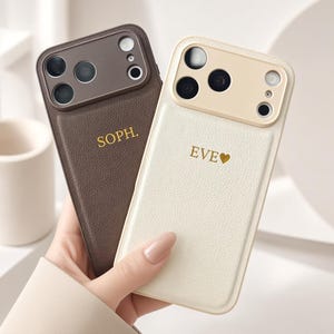 Op de afbeelding: Twee mobiele telefoonhoesjes met een textuurafwerking en een uitsparing voor de camera. De ene is donkerbruin met de gouden tekst SOPH. en de andere is crème met de gouden tekst EVE gevolgd door een hartje. Beide hebben een bijpassende camerarand.