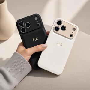 Op de afbeelding: Twee smartphonehoesjes, één zwart en één wit, met een textuurafwerking en een uitsparing voor de camera. Het zwarte hoesje is voorzien van de gouden initialen F.K, terwijl het witte hoesje de gouden initialen A.S bevat. Beide hebben een bijpassend frame.