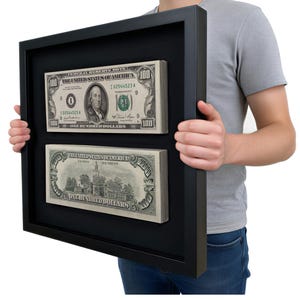 May include: A black shadow box frame containing two stacks of one hundred dollar bills. The top stack displays the front of a bill featuring Benjamin Franklin, while the bottom stack shows the back of a bill depicting Independence Hall.