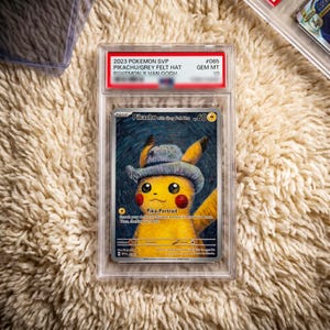 May include: A graded 2023 Pokemon SVP Pikachu with Grey Felt Hat trading card in a clear plastic slab. The card features a yellow Pikachu wearing a blue hat against a blue swirling background, with the text Pikachu with Grey Felt Hat and Pika-Portrait.