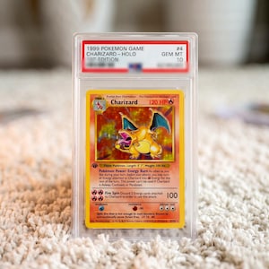 May include: A 1999 Pokemon Game Charizard Holo trading card graded GEM MT 10, encased in a clear protective plastic slab. The card features a yellow border, a fire-type Charizard illustration, 120 HP, and text detailing Energy Burn and Fire Spin.