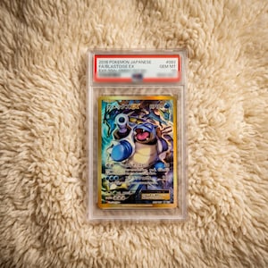 以下が含まれることがあります： 2016年製ポケモンカードゲームの日本語版カメックスEX。GEM MT 10の鑑定済みで、透明なプラスチック製の保護ケースに入っています。カードには、大砲を備えた青いカメックスのキャラクターのカラフルなイラストと、日本語のテキストが描かれています。