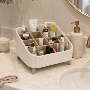 Op de afbeelding: Een crèmekleurige cosmetica-organizer met een geribbelde buitenkant en vier kleine pootjes. De binnenkant is voorzien van meerdere vierkante vakjes gevuld met diverse huidverzorgingsflesjes, tubes en make-up items, waaronder een tube reinigingsmiddel.