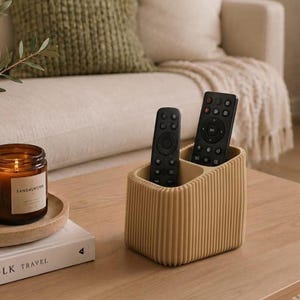 Peut inclure: Un organisateur de rangement rectangulaire beige avec une texture côtelée verticale et deux compartiments intérieurs. Deux télécommandes noires sont placées à la verticale dans l'organisateur, qui présente des coins arrondis et une finition mate.
