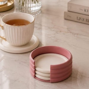 May include: A white ribbed ceramic teacup with a gold-tone handle and rim sits on a matching white saucer. Beside it, a stack of three white circular coasters is held within a dusty rose pink cylindrical holder with a vertical cutout opening.
