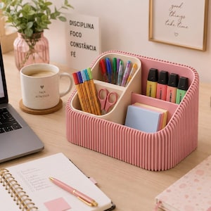 Pode incluir: Um organizador de secretária rosa com exterior canelado e vários compartimentos. O organizador contém várias canetas coloridas, uma pequena tesoura rosa e uma pilha de notas autocolantes em tons pastel. Os divisores internos são creme.