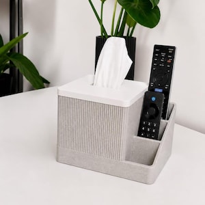 Peut inclure: Un porte-boîte à mouchoirs rectangulaire gris avec un couvercle blanc et une texture nervurée verticale sur les côtés. L'unité dispose d'un compartiment latéral intégré contenant deux télécommandes noires, une longue et une courte, avec des boutons.