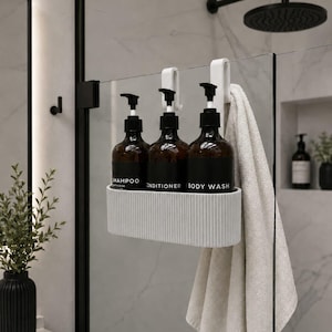 Peut inclure: Un panier de douche gris clair nervuré suspendu à une paroi en verre, contenant trois flacons ambrés avec des pompes noires étiquetés Shampoo, Conditioner et Body Wash, avec une serviette beige drapée sur le côté du panier.