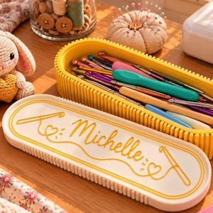 Op de afbeelding: Een gele ovale opbergdoos met een wit deksel waarop de naam Michelle in gele schrijfletters staat, vergezeld van een haaknaaldgrafiek. De doos bevat diverse kleurrijke haaknaalden en staat naast een gehaakt konijntje.