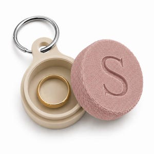 Op de afbeelding: Een beige rond ringdoosje met een zilveren metalen sleutelring. Het doosje is geopend en toont een eenvoudige gouden ring aan de binnenkant. Het deksel is gestructureerd, oudroze en heeft een grote, in reliëf gedrukte hoofdletter S bovenop.
