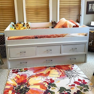 May include: A white wooden daybed featuring three storage drawers along the base and a trundle drawer underneath. The bed is dressed with a patterned orange and brown giraffe-print bedding set, including a pillow and a plush giraffe toy.