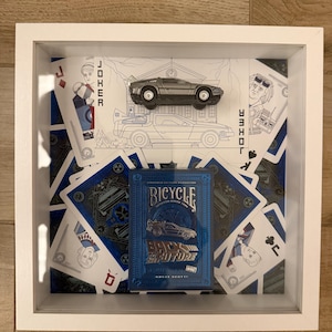 Op de afbeelding: Een witte schaduwdooslijst met een blauw Bicycle Back to the Future kaartspel, een zilveren speelgoedauto en diverse verspreide speelkaarten met illustraties van personages en de DeLorean tijdmachine-auto.
