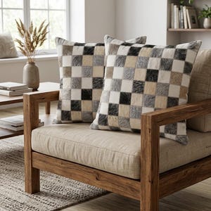 May include: Two square throw pillows featuring a checkered pattern made of patches in shades of black, white, gray, and tan. The pillows have a textured, hair-on-hide appearance and are positioned on a wooden chair with a light beige seat cushion.