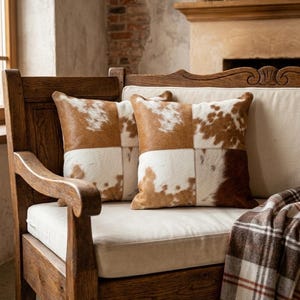 May include: Two square throw cushions featuring a patchwork design of brown and white cowhide leather. The cushions are constructed with visible stitching connecting the hide panels, creating a textured surface with natural hair-on-hide patterns.