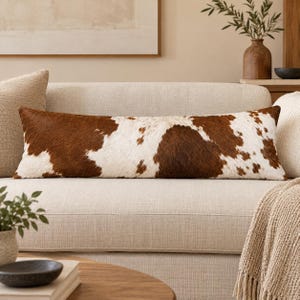 May include: A long, rectangular lumbar pillow featuring a natural cowhide pattern with irregular patches of dark brown and white fur. The pillow has a soft, textured appearance and is positioned horizontally across the center of a light beige sofa.