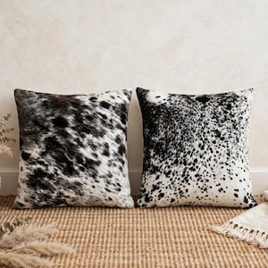 May include: Two square throw pillows featuring a black and white cowhide pattern. The left pillow displays large, irregular black patches on a white base, while the right pillow shows a dense, speckled pattern of small black spots on white.