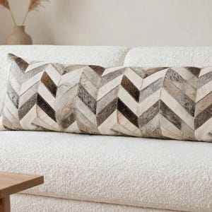 May include: A long rectangular lumbar pillow featuring a chevron pattern made of stitched panels in shades of cream, gray, brown, and black. The pillow is positioned horizontally across the center of a textured cream-colored sofa.